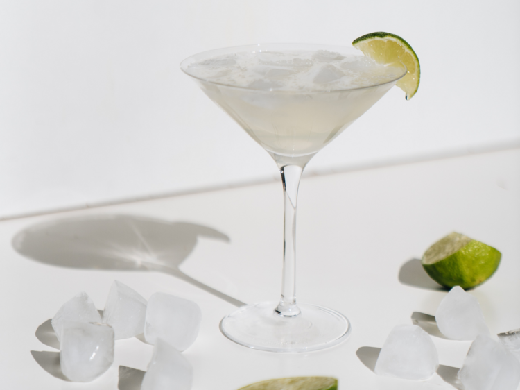 On a white table there is a margarita in a glass next to ice cubes and lime wedges