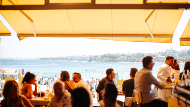 People at tables, with Bondi Beach and the ocean in the background.