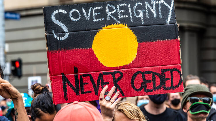 A protestor holding a sign painted like the Aboriginal flag, reading 'sovereignty never ceded'.