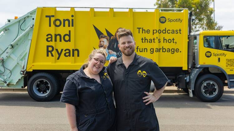 Podcasters Toni and Ryan standing in front of a yellow garbage truck.