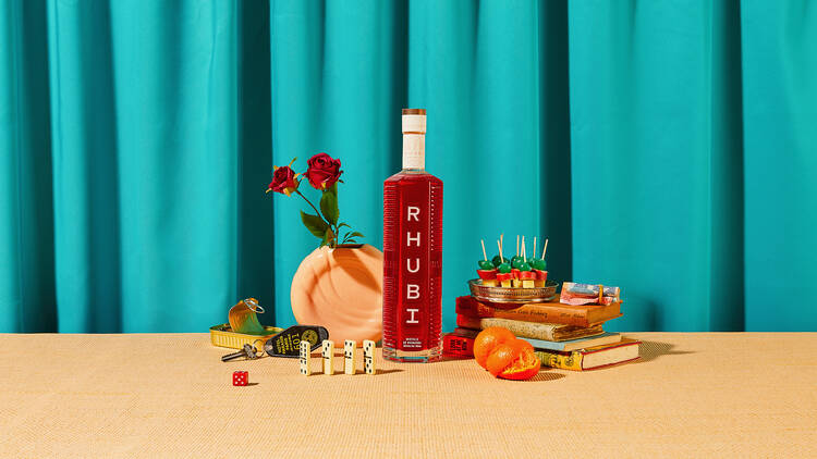 A bottle of Rhubi with some roses, books and snacks in front of a blue curtain