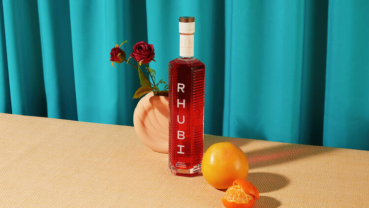 A bottle of Rhubi in front of a blue curtain, with oranges and roses in a pink vase