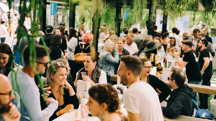 A crowd of people talk animatedly and drink gin at Juniperlooza, under lush hanging plants.
