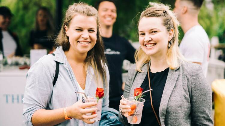 Two women wearing casual clothes hold gin and tonics with roses as garnish, while smiling at the camera.