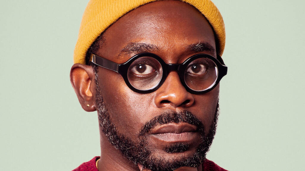 Close-up photograph of playwright James Ijames wearing a yellow beanie and black plastic-framed glasses.