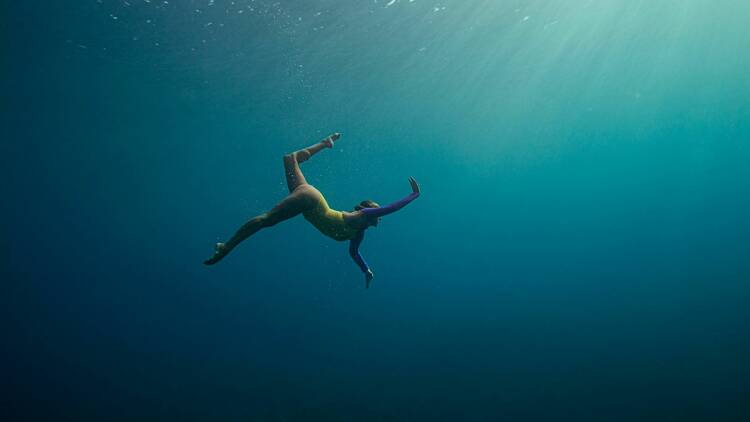 In deep blue water streaked by light from the surface above, a woman is balletically stretching her arms and legs underwater.