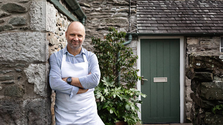 Chef Simon Rogan standing outside his restaurant wearing a blue shirt and white apron