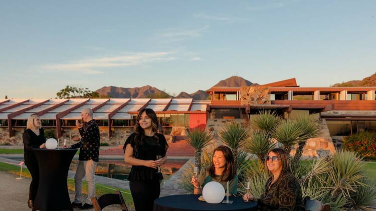 A handful of people stand and sit with glasses of wine as the shadows of sunset fall around them. In the background is the triangular lawn and home of Taliesin West.