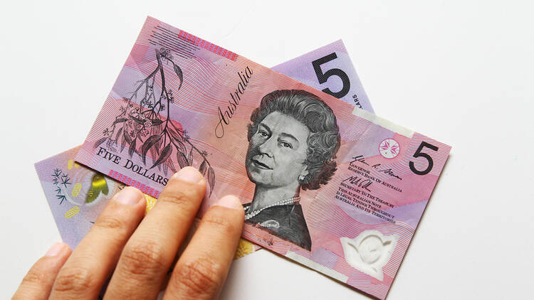 A person holds two purple five dollar notes with an image of the queen on it