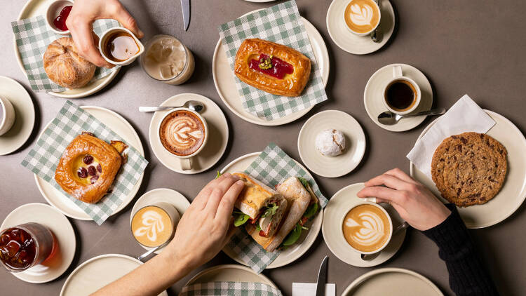 On a grey table there is a selection of coffees pastries and sandwiches on white plates with green and white chequered paper