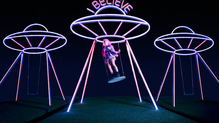 Three neon signs shaped like space ships with a person swinging from one.