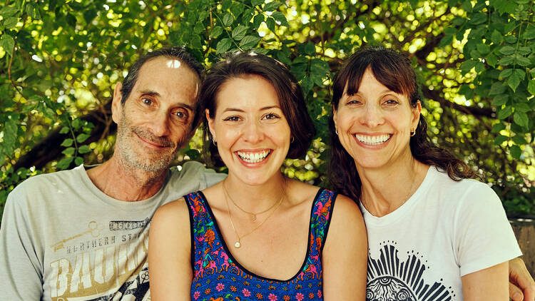 In front of a green tree there are two parents sitting on either side of their adult daughter smiling for the camera