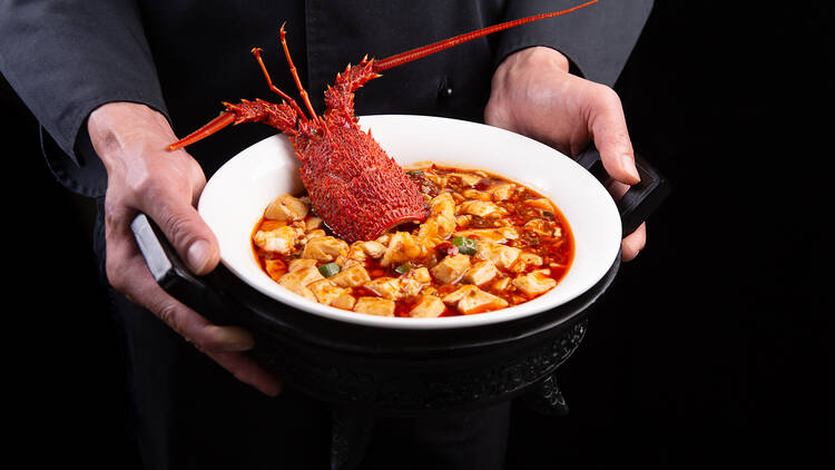Someone holds a white bowl filled with tofu and lobster with a lobster head used as decoration