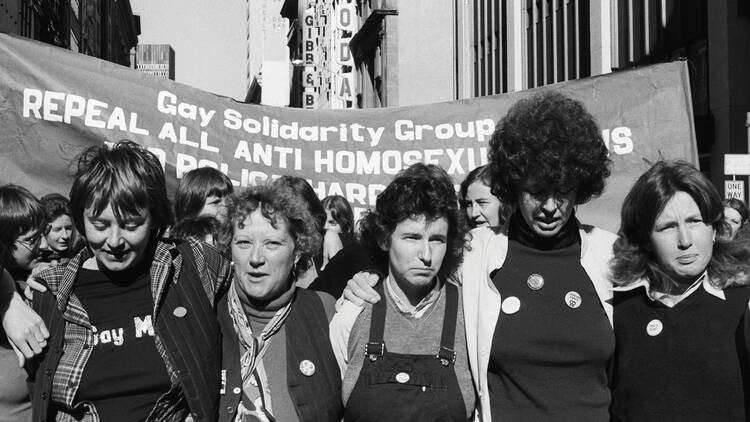 A black and white photo of women in a protest, with a sign behind them that says: Gay Solidarity Group