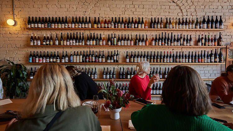 On a brick wall there are shelves of wine bottles and in front there is a large communal table with people sitting around drinking wine