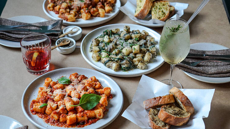 On a table there are three large plates of gnocchi next to garlic bread and several cocktails