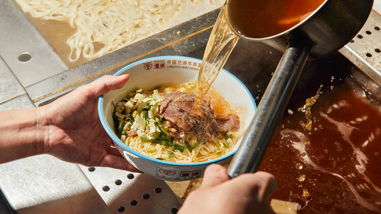 Someone pours broth into a bowl of beef noodles