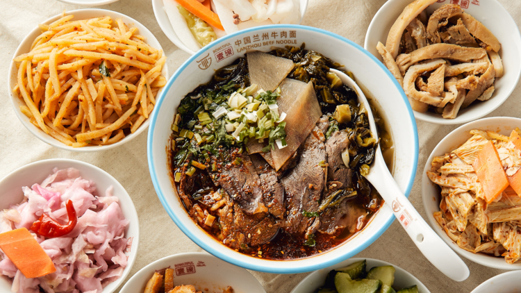 There is a bowl of beef noodles next to many small bowls of side dishes includes pickles vegetables and meats