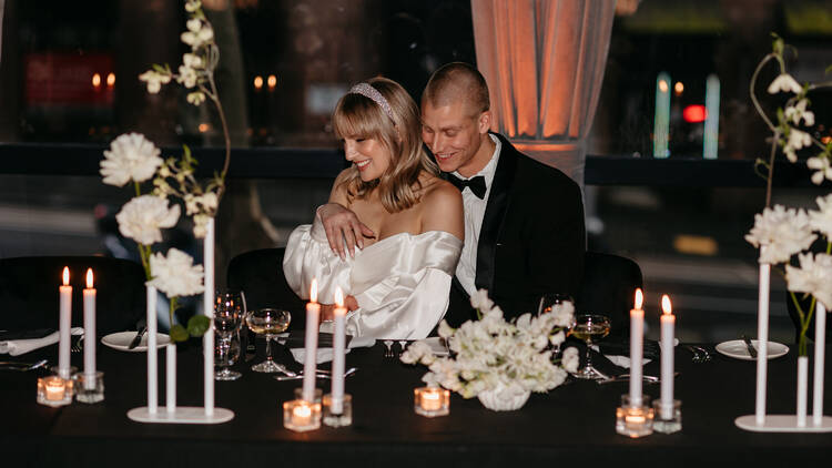 A bride and groom cuddle at a table filled with candles and flowers.