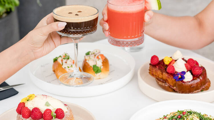 Two people with an Espresso Martini and a Watermelon Margarita cheers over a table with plates of food.