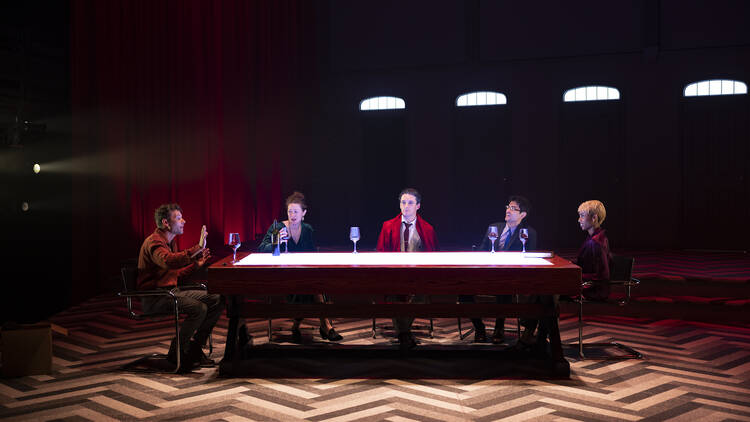 Five people sit around a table on a stage drinking wine.