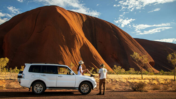 A white truck is parked at the base of Uluru with one person sitting on the hood on the car and one standing next to it