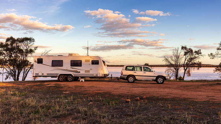 A four-wheel drive tows a caravan along a dirt track by a lake.