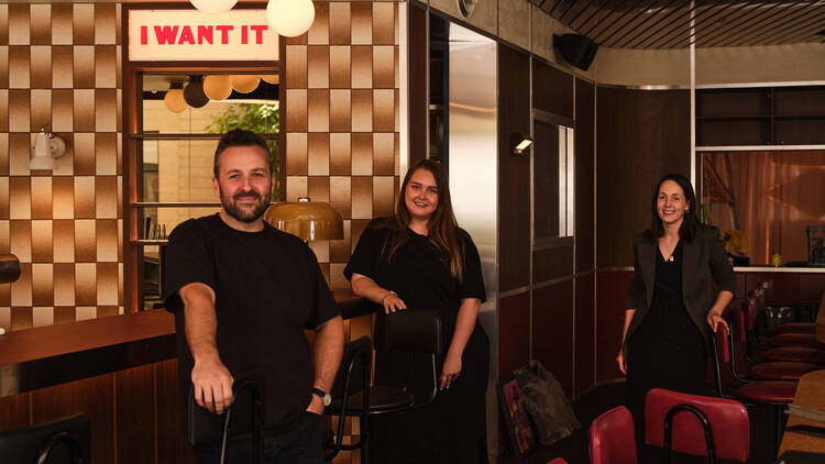 Three people standing in a restaurant, with a sign saying I WANT IT above them.