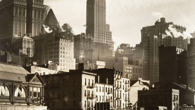 A black-and-white photo by Berenice Abbott showing West Street in 1936 with a few skyscrapers and several vehicles.