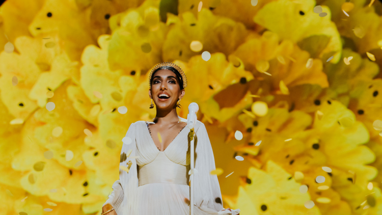 Poet Rupi Kaur smiles in a gold shower against a yellow backdrop