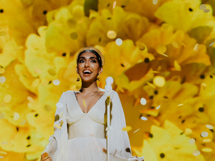 Poet Rupi Kaur smiles in a gold shower against a yellow backdrop