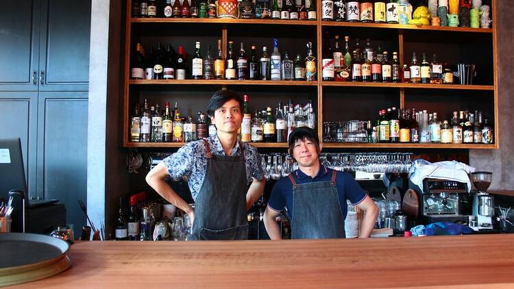 Two staff behind the bar, with shelves full of Japanese alcohol.