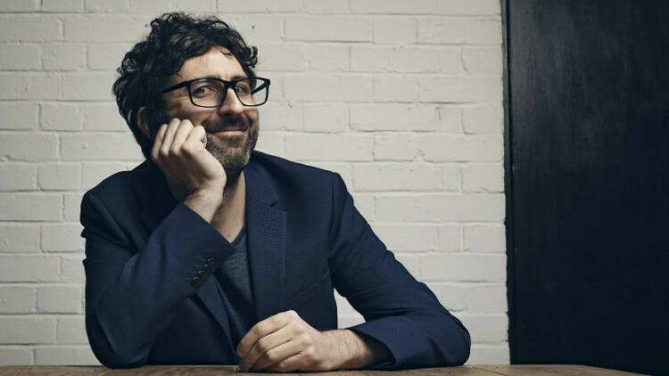 Comedian Mark Watson sitting at a table with his face in his hands.