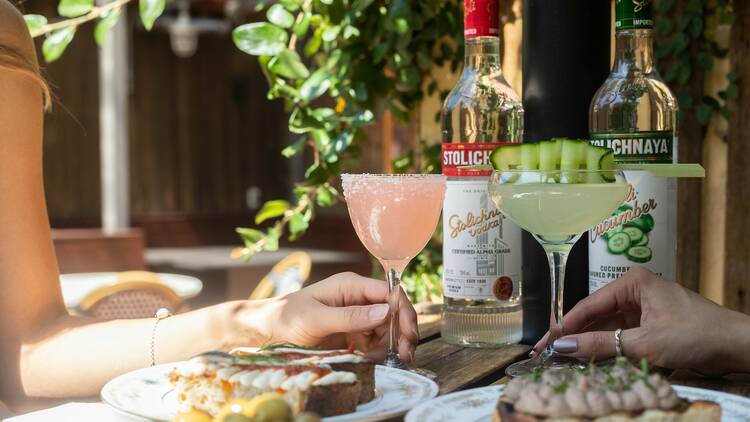 Two hands hold a pink sugar rimmed Martini and a green martini garnished with cucumber.