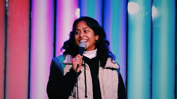 Comedian Sureni Weerasekera holds a microphone in front of a colorful background.