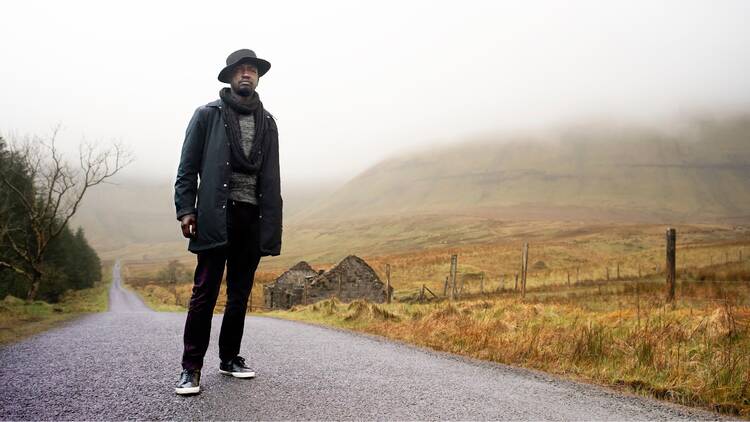 A man named Paddy Hazelton stands on an empty street in what appears to be the Irish countryside. 