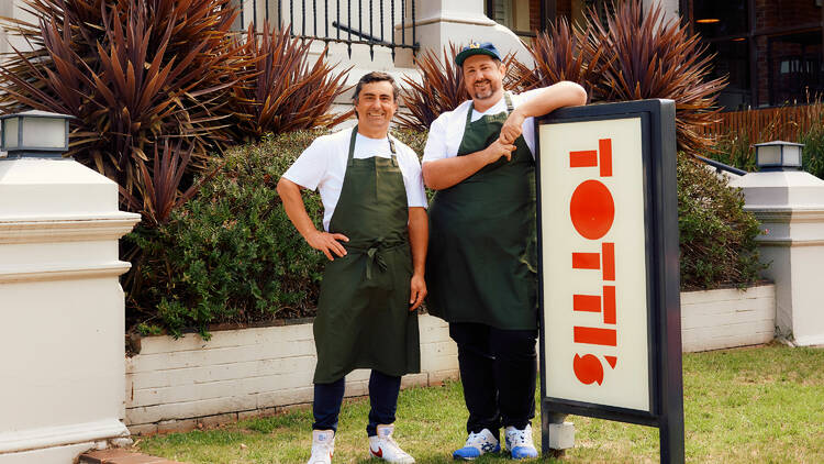 Two men in chef's aprons lean on a sign that says 'Totti's'.