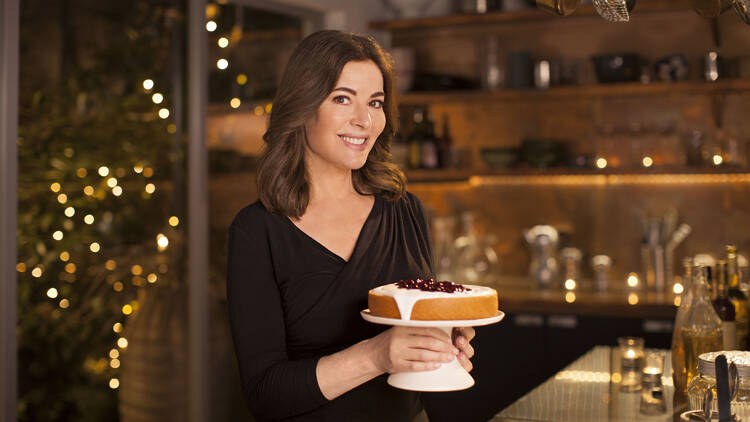 Nigella Lawson holding a cake with icing and berries on it in a cosy kitchen.
