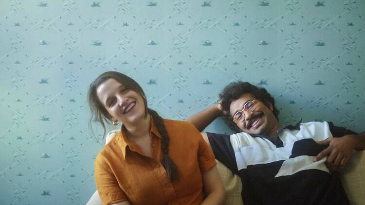 A man and woman sit on a couch smiling in front of a blue wall. 