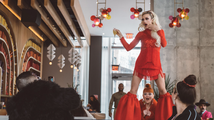A performer in a red dress stands on a bar holding a drink.