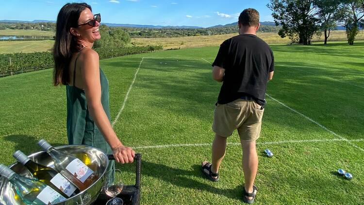 A man and a woman stand on a boules green, playing next to a wine bucket filled with various bottles.