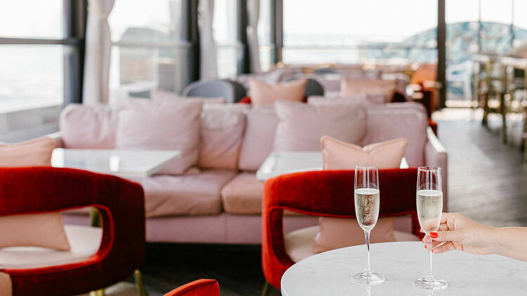 A bar with a pastel pink lounge and red chairs, with 2 glasses of Champagne in the foreground.