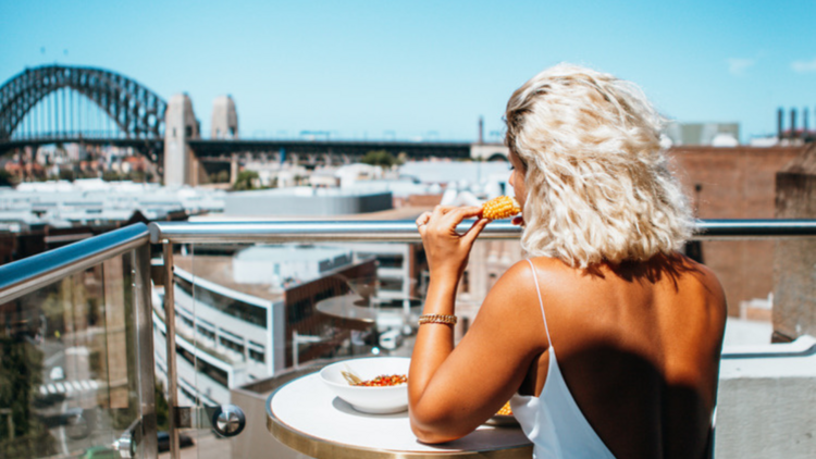 A woman with a white, backless dress and blonde hair eating something on a rooftop bar balcony, looking at a view of the Sydney Harbour Bridge.