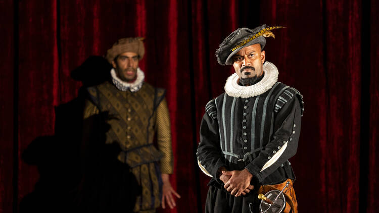 Two men in Victorian-era costumes stand on a stage in front of a red carpet.