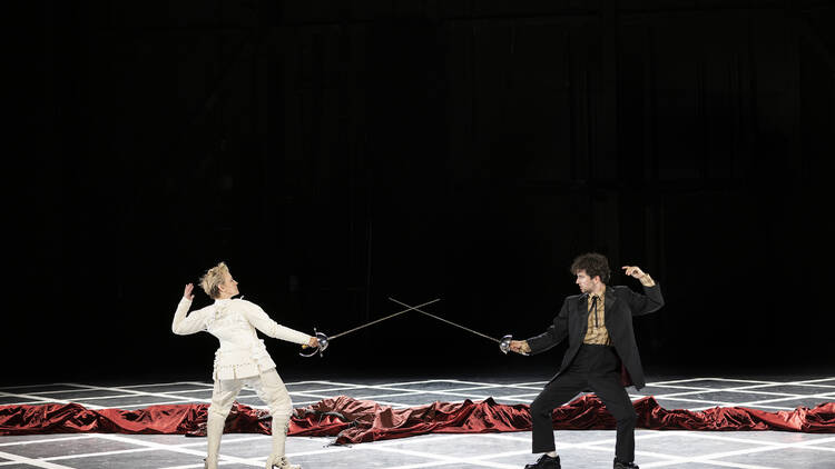 Two actors (one in all-white, one in all-black) sword fight on an empty stage.