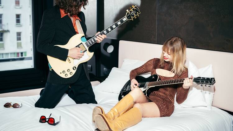 A man and a woman playing electric guitars on a hotel bed