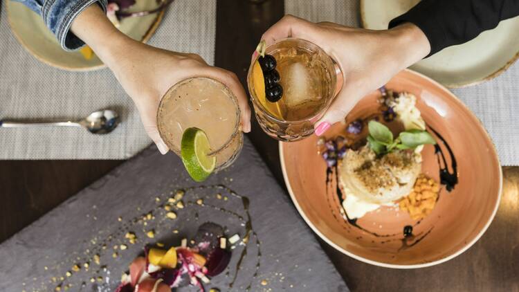 Two people toasting cocktails over plates of food