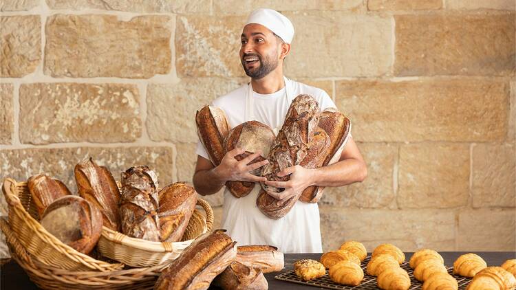 A baker with bread and croissants against a sandstone wall