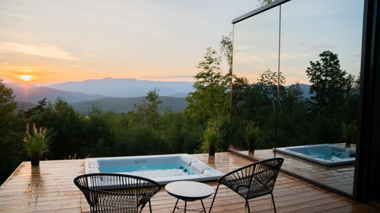 With a sunset over the mountains in the distance, one wall of the glass house is visible next to the patio with embedded hot tub