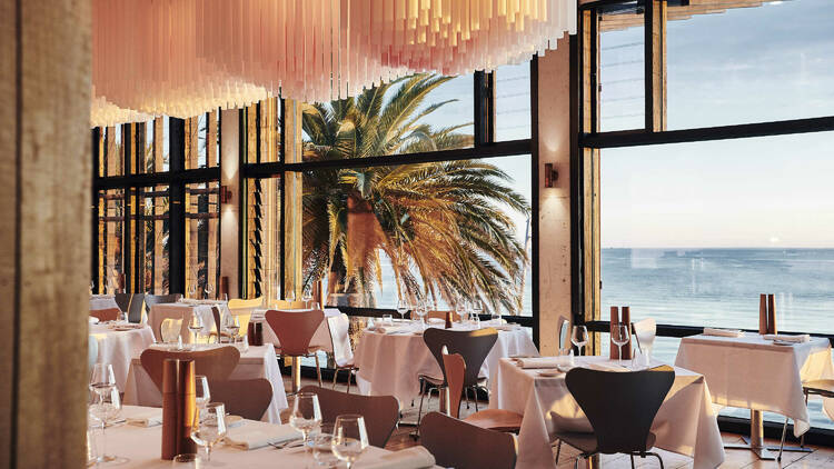 A light dining space overlooking the ocean with floor-to-ceiling windows, modern chandeliers and dining tables.